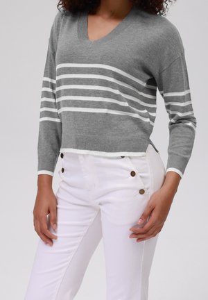 Vrouw draagt een grijze V-hals trui met witte horizontale strepen en witte broek met hoge taille met bruine knopen op de heupen.