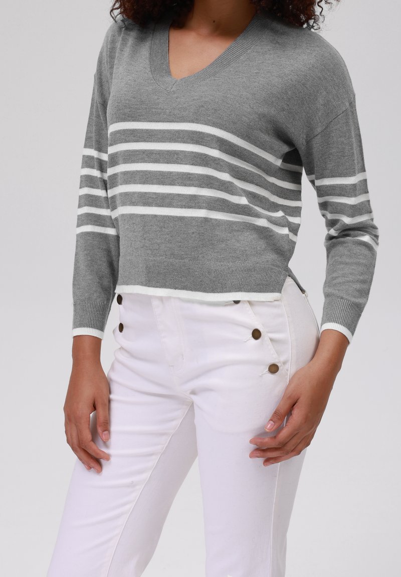 Femme portant un pull gris en V avec des rayures blanches horizontales et un pantalon blanc taille haute avec des boutons marron sur les hanches.