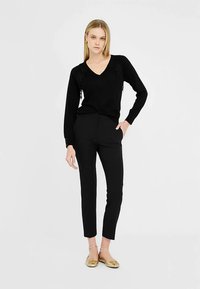 Sort v-hals sweater med blomstret blonde detaljer, kombineret med sorte slim-fit bukser og guld flade sko. Glat stof, skræddersyet design og tætsiddende silhuet.