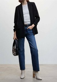 Blazer nero sartoriale su una maglietta grigia chiara, abbinato a jeans blu dritti. Stivaletti bianchi con tacco e una borsa con motivo completano il look.