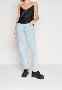 Camisole en satin noir à fines bretelles, jean bleu clair droit, et bottes noires à semelles épaisses. Fond uni.