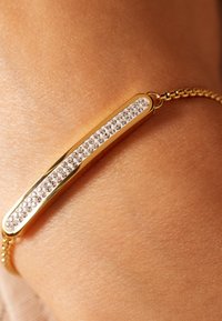 Gouden armband met een slanke staaf waarop een rij heldere strass-stenen, een getextureerde ketting en een gladde, gepolijste oppervlakte.