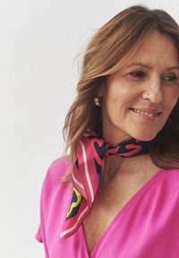 Silk tørklæde i pink med et geometrisk mønster, der har hjerter, striber og klare farver. Båret knyttet om halsen, der komplimenterer en pink top.