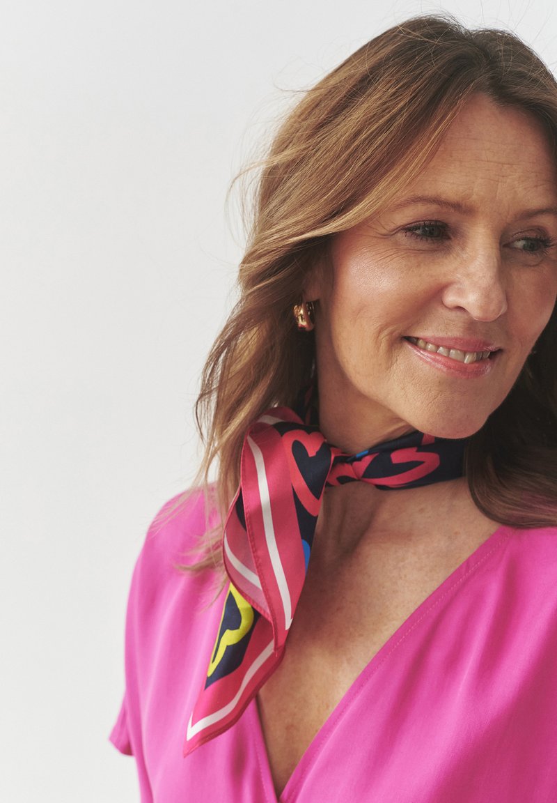 Silk tørklæde i pink med et geometrisk mønster, der har hjerter, striber og klare farver. Båret knyttet om halsen, der komplimenterer en pink top.