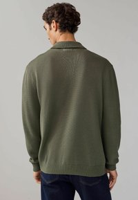 Pulover verde tricotat cu un design cu guler, manșete zimțate și un finisaj texturat, purtat cu blugi închiși. Vedere din spate.