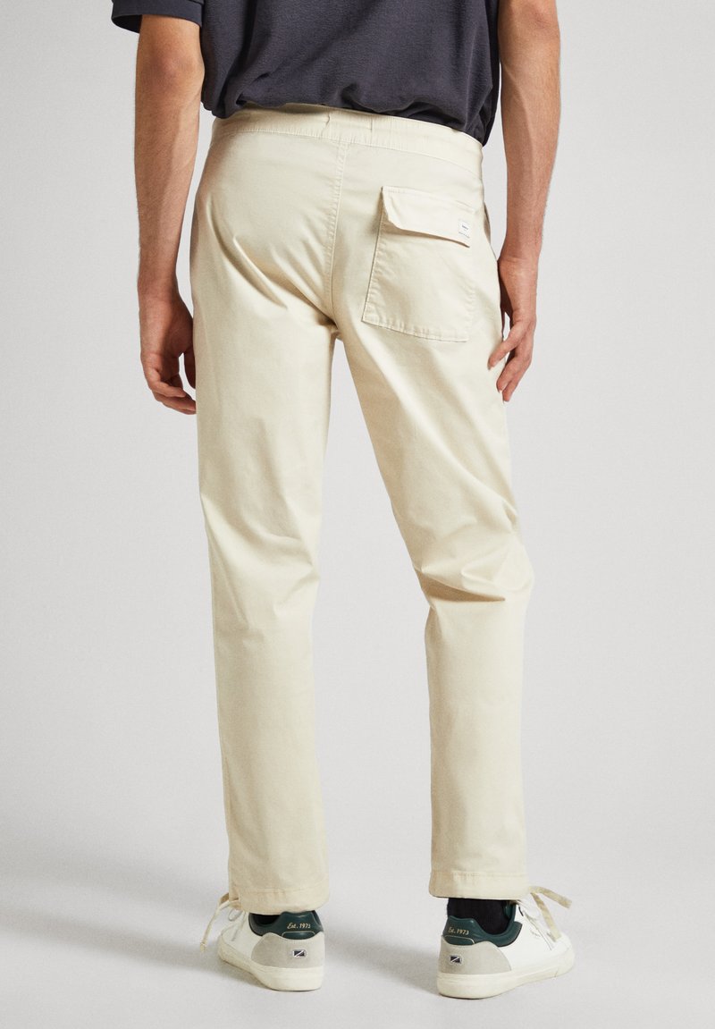 Hombre que lleva pantalones beige de corte recto con bolsillo trasero con solapa y zapatillas blancas con detalles en verde oscuro, de pie frente a un fondo liso.