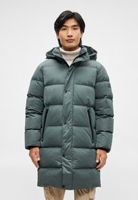 Pitkä, vihreä puhvitakki huppulla, jossa on tikatut osat, mustat vetoketjulliset taskut ja napillinen etuosa. Tekstuuri tuo lisää yksityiskohtia.