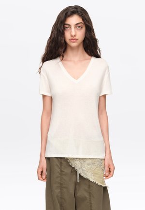Joven mujer con cabello largo y ondulado que lleva una camiseta blanca con cuello en V y pantalones oliva con un parche decorativo de tela beige.