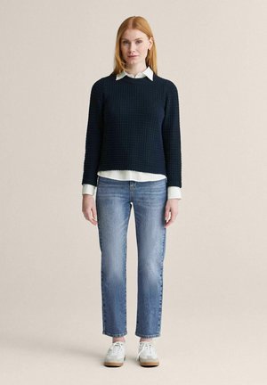 Kvinde med glat rødt hår iført en sort tekstureret sweater over en hvid skjorte, blå jeans og hvide sneakers, stående mod en neutral baggrund.