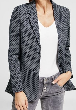 Vrouw draagt een zwart-grijs patroonblazer over een wit V-hals T-shirt, gecombineerd met grijze jeans met knoopsluiting, hand in de zak.