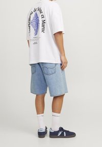 Vit oversized t-shirt med lila grafik och text på baksidan, tillsammans med ljusblå denimshorts och marinblå fritidsskor med vita strumpor.