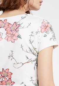 Bílý krátký rukáv T-shirt s květinovým potiskem, který zobrazuje růžové květiny a zelené listy, vykazující hladkou texturu a ležérní design.