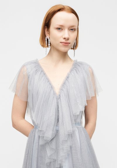 Φόρεμα ανοιχτού γκρι με στρωματοποιημένα, διάφανα μανίκια από τούλι και βαθύ ντεκολτέ σε σχήμα V. Λαμπερή υφή, εφαρμοστή μέση και τσέπες σε κάθε πλευρά.