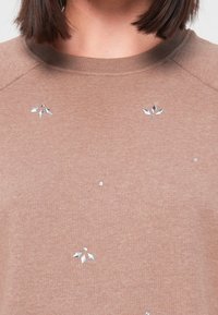 Pull marron à col rond avec de petits clous argentés en forme de feuille et ronds dispersés à l'avant, porté par une personne aux cheveux courts et foncés.