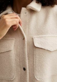 Chaleco de lana beige con cuello, delante con tapeta, dos bolsillos en el pecho y broches a presión. La tela tiene un acabado suave y texturizado.