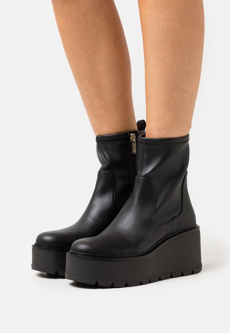 Bottes à plateforme en cuir noir à fermeture éclair latérale portées sur des jambes nues sur un fond uni.