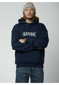 Sudadera con capucha de color azul marino con "Kaporal Jeans" impreso en blanco. Cuenta con un bolsillo tipo canguro y puños de canalé. Combinada con jeans de mezclilla.