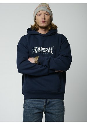Sudadera con capucha de color azul marino con "Kaporal Jeans" impreso en blanco. Cuenta con un bolsillo tipo canguro y puños de canalé. Combinada con jeans de mezclilla.