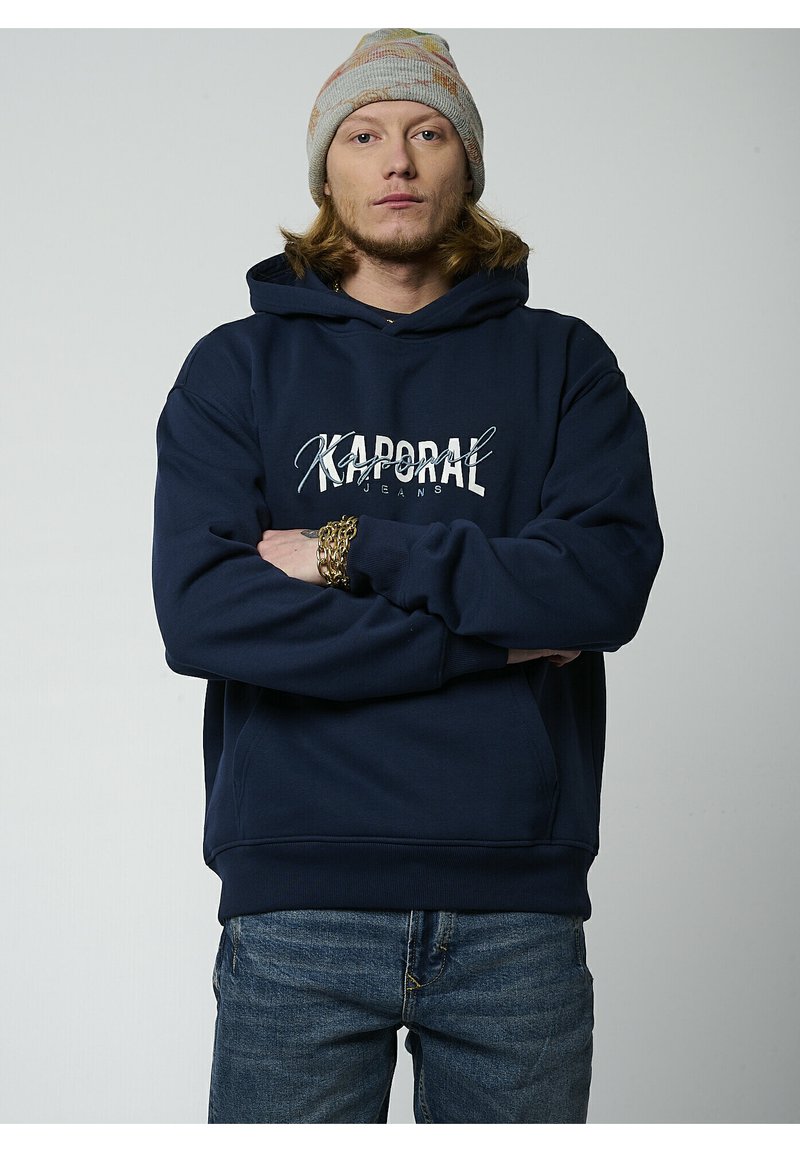 Sudadera con capucha de color azul marino con "Kaporal Jeans" impreso en blanco. Cuenta con un bolsillo tipo canguro y puños de canalé. Combinada con jeans de mezclilla.