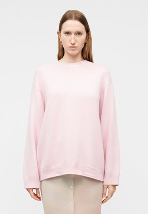 Νεαρή γυναίκα με ίσια ανοιχτό καστανά μαλλιά φοράει ένα φαρδύ ανοιχτό ροζ πουλόβερ και μπεζ φούστα, στέκεται μπροστά σε ένα απλό ανοιχτό φόντο.