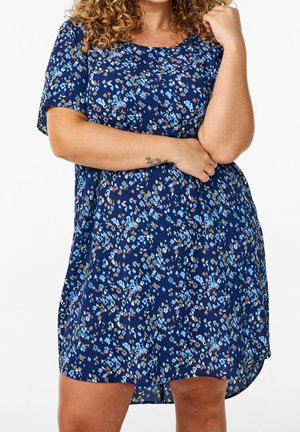 Femme portant une robe bleue à manches courtes avec un petit imprimé floral blanc et marron, bras croisés, montrant un tatouage au poignet.