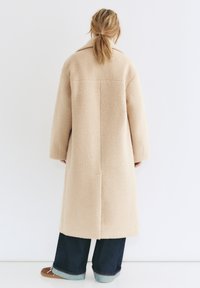 Beżowy wełniany płaszcz o luźnym kroju, z dużym kołnierzem i długimi rękawami, w połączeniu z ciemnoniebieskimi dżinsami i brązowymi butami. Teksturowana, ciepła tkanina.
