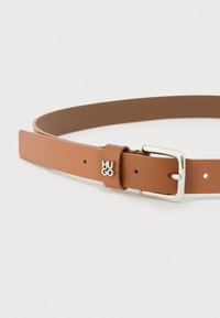 Kremeni kožni remen sa srebrnom pravokutnom kopčom i metalnim logotipom "HUGO" na petlji, položen ravno na bijeloj pozadini.