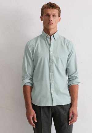 Camicia button-up verde chiaro con colletto, taschino singolo sul petto, maniche arrotolate e una texture liscia, abbinata a pantaloni scuri.