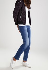 Černá vzorovaná bunda se stříbrnými zipy, bílým tričkem, modrými džíny a bílými slip-on botami. Bunda má kapuci a žebrované manžety.