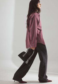 Gestreiftes karminrotes Hemd mit weiten Ärmeln, kombiniert mit schwarzen weiten Jeans und spitzen burgunderfarbenen Pumps. Kleine schwarze Handtasche mit einem gemusterten Trageriemen.