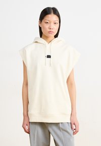 Helle beige ärmellose Kapuzenjacke mit Kordelzug, lockerer Passform und einem kleinen schwarzen Logo-Detail auf der Brust. Weiches Baumwoll-Mischgewebe.