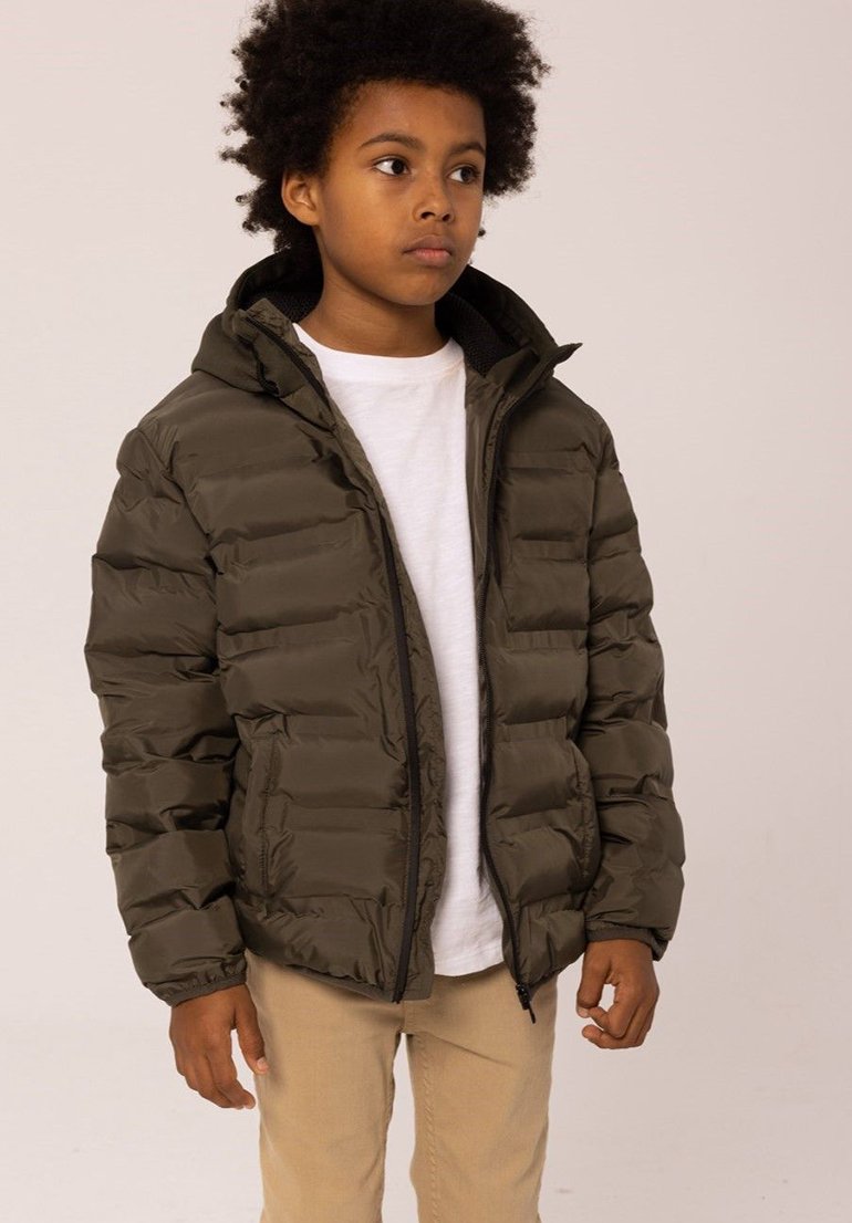 Giubbotto imbottito verde oliva con cappuccio, texture trapuntata, chiusura frontale con zip e polsini elasticizzati, indossato sopra una t-shirt bianca e pantaloni beige.