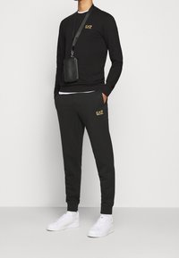 Camisa negra de manga larga con logotipo amarillo, combinada con pantalones de chándal gris oscuro que presentan un logotipo a juego, complementada con zapatillas blancas y una bolsa negra.