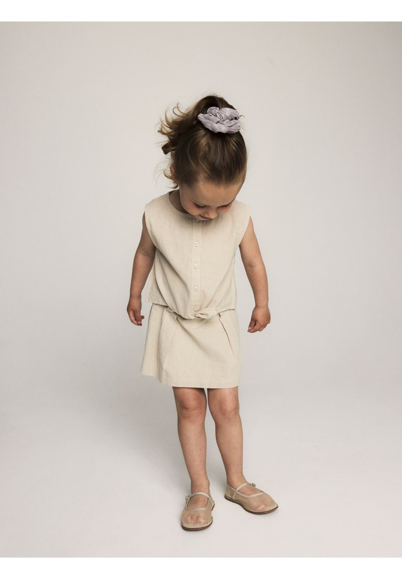 Robe sans manches beige avec un détail de taille nouée, deux poches avant et des boutons. Associée à des chaussures transparentes et un chouchou texturé pour les cheveux.