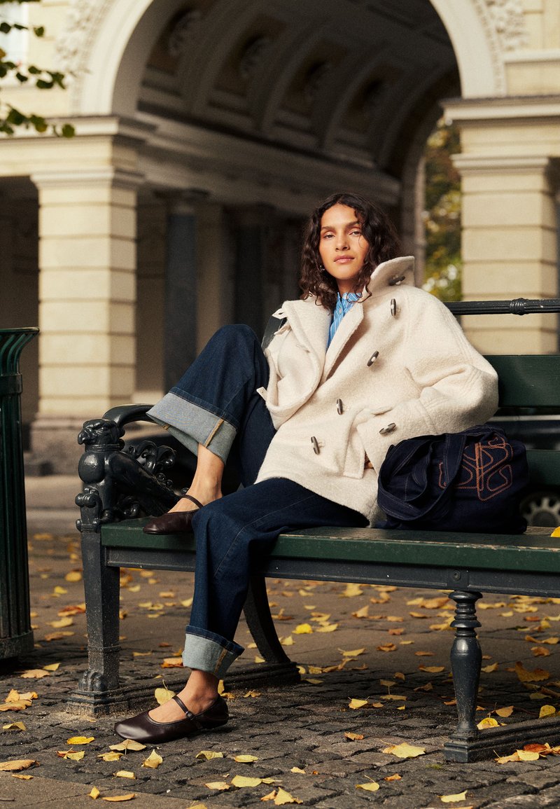 Femme aux cheveux bouclés assise sur un banc vert en plein air, portant un manteau blanc, un jean bleu et des chaussures marron, avec un sac sombre à côté d'elle.