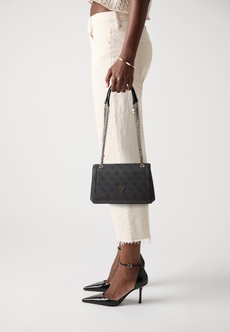 Bolso de mano negro texturizado con asas de cadena, que presenta un logo triangular. Combinado con pantalones cortos color crema y tacones negros de punta.