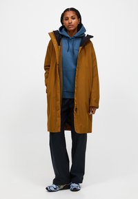 Bruine waterdichte jas met een capuchon, ritsluiting en zijzakken; gedragen over een blauwe hoodie en zwarte broek; heeft met patroons versierde schoenen.