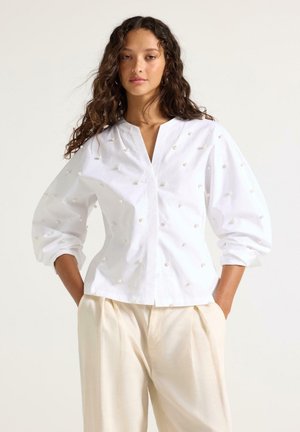 Nainen, joka pukeutunut valkoiseen helmirakein koristeltuun paitaan ja kermaisiin leveälahkeisiin housuihin, seisoo käsine taskuissa yksinkertaisen taustan edessä.