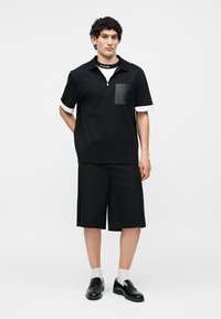 Άνδρας που στέκεται φορώντας μαύρο κοντομάνικο πουκάμισο με δερμάτινη τσέπη, μαύρες μακριές βερμούδες, άσπρες κάλτσες και μαύρα λοφάρια σε λευκό φόντο.