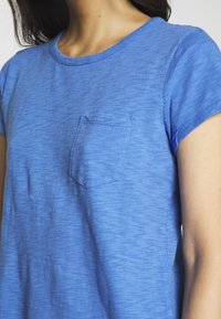 Blå bomullströja med rund halsringning och korta ärmar, med en liten bröstficka. Texturen verkar mjuk och lätt.