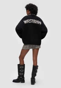 Przesadny czarny sweter robiony na drutach z białym napisem "WRSTBHVR" z tyłu, w połączeniu z teksturowaną szaryą spódnicą i czarnymi, wysokimi bucikami z klamrami.