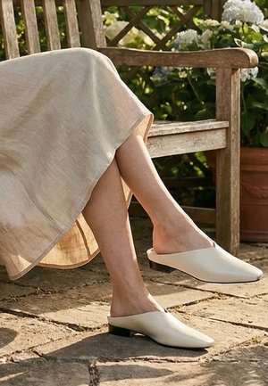 Osoba sedící venku na dřevěné lavici se zkříženýma nohama, oblečená v béžové sukni a bílých slip-on plochých botách na kamenné terase.