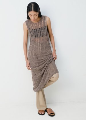 Γυναίκα που φοράει αμάνικο, καφέ φόρεμα με υφή πάνω από μπεζ παντελόνι, σηκώνοντας το τελείωμα του φορέματος, στέκεται μπροστά σε έναν λευκό τοίχο με το κεφάλι ελαφρώς σκυμμένο.