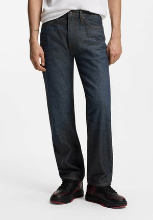 Mann trägt dunkelblaue Jeans mit geradem Bein, weißes eingestecktes Hemd und schwarze Sneakers mit roten Akzenten, steht vor weißem Hintergrund.