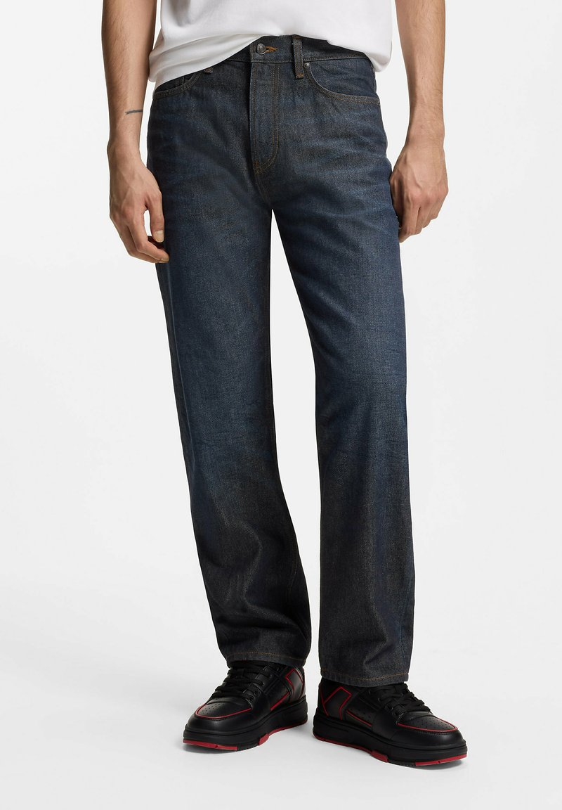 Homme portant un jean droit bleu foncé, une chemise blanche rentrée, et des baskets noires avec des touches rouges, debout sur fond blanc.