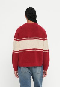 Persona con capelli intrecciati che indossa un maglione rosso con strisce bianche sulla schiena e jeans blu, in piedi di fronte a uno sfondo bianco semplice.