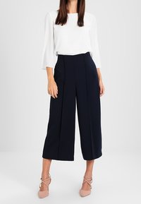 Marineblaue weit geschnittene Hose mit hoher Taille, plissiertem Vorderteil und glattem Stoff. Kombiniert mit einem weißen Oberteil und hellrosa Riemchen-Pumps.