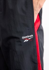 Reebok tepláky z čierneho materiálu s červenými akcentmi. Obsahujú vyšívané logo a bočné vrecká. Hladký povrch a ľahký dizajn.