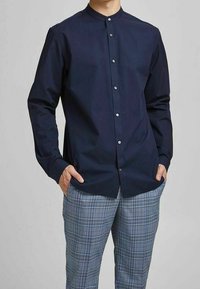 Camicia a maniche lunghe blu navy con colletto alla coreana, dotata di bottoni bianchi e vestibilità slim. Abbinata a pantaloni scozzesi azzurri chiari.