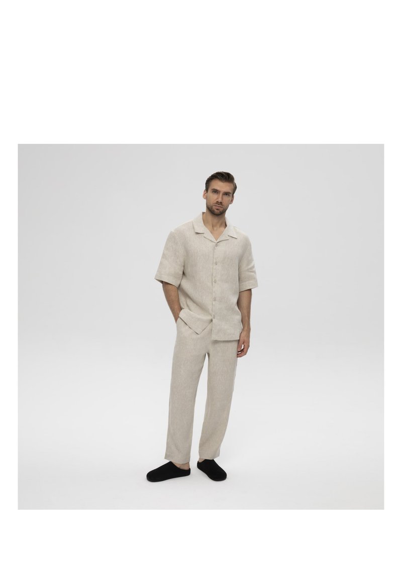 Mann steht mit den Händen in den Taschen, trägt ein hellbeiges kurzärmeliges Hemd mit Knöpfen, passende Hose und schwarze Slipper.